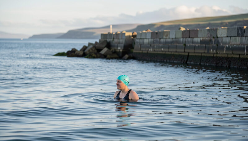 A lone swimmer in a bright swim cap standing waist deep in cold blue sea beside a stone breakwater on a Scottish coast